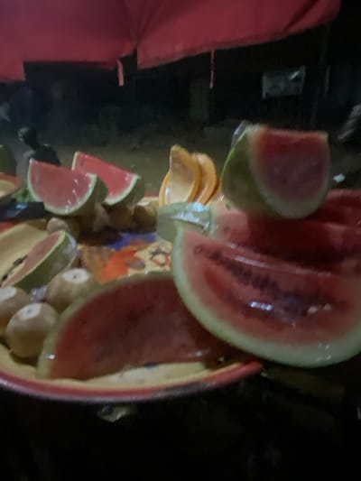 A vibrant assortment of sliced watermelons and peeled kiwis fill a large bowl. Chunks of watermelon, ranging from large wedges to smaller triangular pieces, are prominently displayed. The kiwis, appearing round and light brown, nestle among the watermelon slices. The bowl itself, with a colorful artistic pattern, adds to the visually appealing arrangement. The setting appears to be dimly lit, with a pink umbrella and dark backgrounds.