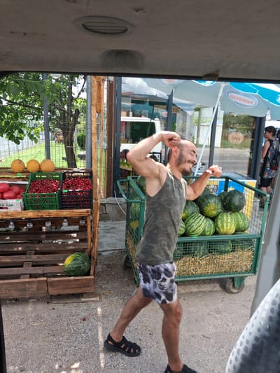 A man with a shaved head and beard, wearing a camouflage tank top and skull-patterned shorts, poses with his arms raised and fists clenched. He stands in front of a cart filled with watermelons. To his left, a wooden display holds various fruits, including cherries in green and black crates, and several round, yellow fruits, possibly melons. A watermelon rests on a wooden pallet to the left of the fruit display. In the background, a building with blue umbrellas and a sign that says "OPEN" is visible. The scene appears to be an outdoor market. The image was taken in Kukorevo, Bulgaria.