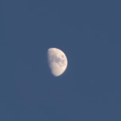 A half-illuminated moon is visible against a clear, dark blue sky. The moon is off-center, positioned in the lower right portion of the frame. The illuminated portion of the moon exhibits textured cloud-like formations or craters. The rest of the moon is in shadow, creating a crescent shape. The image is taken in Mas Aliu, Spain.
