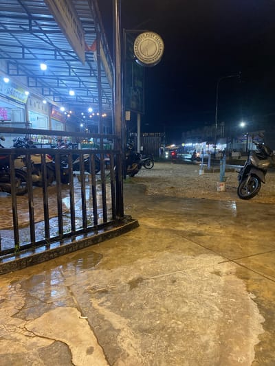 This night-time image depicts a wet street or pathway in an urban or semi-urban setting, likely after rainfall. The perspective is from a slightly elevated position, looking down towards a street lined with shops or stalls.
In the immediate foreground, a dark metal gate or railing extends diagonally across the lower left side of the frame. The concrete ground is visibly wet, showing numerous puddles that reflect the overhead lights. The ground is textured with cracks and various patterns, including what appears to be footprints and debris.
Beyond the gate, several motorcycles, primarily scooters, are parked. A prominent dark grey scooter is positioned on the wet ground in the mid-ground. The area under a large, possibly corrugated metal, awning or roof structure is brightly illuminated by numerous artificial lights. This area appears to house small businesses or market stalls.
Several signs and banners are visible:
- A circular sign, prominently displayed, reads "DOCTOR PENY CLINIC" and below it, "JL. KEUDE GEUDONG ACEH TIMUR", providing a clear location cue for Aceh Timur, Indonesia.
- Other banners are partially visible, with text like "PHARMACY" and "AL-BAROKAH".
- Some signs bear the acronym "BSI", likely referring to Bank Syariah Indonesia.
The sky is dark, indicating it is nighttime. A bright streetlight is visible in the upper right, casting light into the dark sky. Faint lights from other establishments or vehicles can be seen in the distance along the street. No specific individuals are clearly discernible or engaged in any particular activity, suggesting a quiet moment. The wet ground and strong artificial lighting are notable features of the scene.