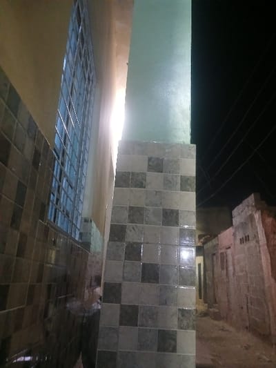 A low-angle shot captures a scene with tiled surfaces. Square tiles in muted tones cover a vertical wall section and also form a reflective, wet-looking floor. A section of glass tiles provides a glimpse into an illuminated space. Above, a concrete structure overhangs the tiled wall, contrasting with the dark sky punctuated by lines, possibly wires. The overall lighting is dim, with a bright spot illuminating the space behind the glass tiles.