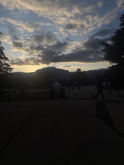 The image captures an expansive outdoor scene during what appears to be the golden hour or sunset, likely in Saint-Martin-d'Hères, France. The perspective views across a paved area towards a distant mountain range under a dramatic sky.
The sky is a prominent feature, filled with a variety of clouds. Layers of grey and white clouds stretch across the upper and middle portions, while closer to the horizon, the setting sun casts a warm, golden-yellow glow on the undersides of the clouds and directly behind the mountains. This strong backlighting creates striking silhouettes of the foreground and mid-ground elements.
In the mid-ground, a majestic mountain range forms a dark, rugged silhouette against the luminous sky, with some higher peaks subtly catching the light, hinting at snow or bright rock faces. Below the mountains, indistinct architectural structures are visible as dark shapes.
The foreground and mid-ground primarily consist of a paved urban or campus-like environment. Several human figures are present, mostly as dark silhouettes. One notable figure walks with purpose on the paved area, appearing to carry a backpack. Other smaller figures are scattered across the plaza. Dark foliage, possibly trees or bushes, frames the scene from the left and top, further emphasizing the dramatic lighting. A small white tent-like structure can also be seen on the paved surface. The activity depicted is general pedestrian movement, suggesting a routine evening scene.
The low angle of the sun creates long shadows extending towards the viewer, enhancing the sense of late afternoon. The combination of the dramatic clouds, the silhouetted mountains, and the warm glow of the sun creates a serene yet dynamic atmosphere. No visible text is discernible in the image.