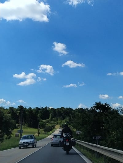 The image depicts a sunny day on a road in Strunkovice nad Volyňkou, Czechia. A motorcycle with a rider wearing a helmet and a black jacket is in the foreground, traveling on a paved road. The motorcycle has two saddlebags and a license plate that reads "BP 5224". Behind the motorcycle, a silver car is visible. Further ahead, other vehicles are present on the road, including a silver Citroën with license plate "4C9 4411" and a white van. The road is bordered by a guardrail and a grassy shoulder. Dense green trees line the right side of the road, forming a wooded hillside. On the left side of the road, there are hay bales and utility poles. The sky is a bright blue with scattered white clouds. A signpost with the text "STRUNKOVICE" is visible on the right side of the road. Another signpost displays "4-246P".