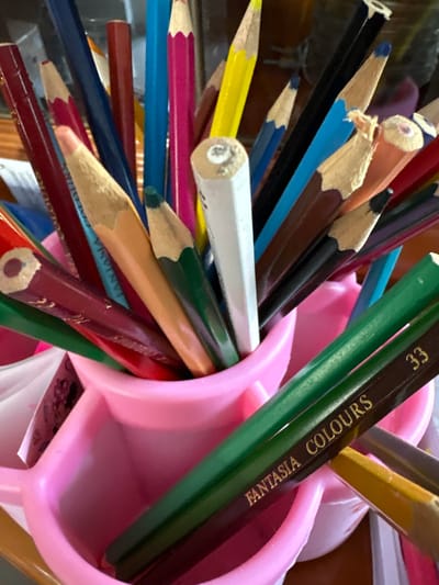 The image shows a collection of colored pencils in a pink pencil holder. The pencils are of various colors, including red, blue, pink, yellow, green, and brown. Some of the pencils have writing on them, such as "FANTASIA COLOURS" and "33". The pencil holder is plastic and has a pink, heart-shaped design. The background is blurred but appears to be a wooden surface, possibly a desk. The image is taken from a close-up perspective, focusing on the pencils and the holder. There are no people or animals visible in the image. The lighting is bright, suggesting it might be daytime or under artificial light. The overall scene depicts a workspace or a child's art supplies. The location of Delbrück, Germany is not discernible from the image itself.