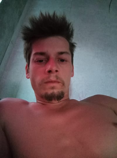 The image is a close-up, low-angle selfie of a shirtless young adult male with spiky brown hair and a goatee. The man's face is illuminated by a red light, casting a reddish hue over his skin. His eyes are open, and he is looking towards the camera. His shoulders and upper chest are visible, also with a reddish tint. The background appears to be a light blue, textured wall, possibly in a room. There are no other people or objects clearly discernible in the scene. The location of Zīlāni, Latvia is not determinable from the visual information present in the image.