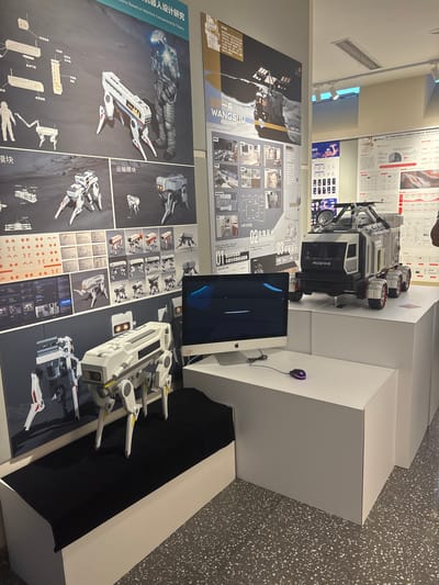 This image depicts an exhibition of robot designs, likely at a museum or research facility, within Minhang District, China. The foreground features two white, four-legged robot models positioned on a black surface and a white pedestal. A computer monitor displaying a blue graphic and a purple-lit mouse are situated on a white table to the left of the robot models. Dominating the background is a large display poster showcasing various robot designs, including a robotic dog, a quadrupedal robot, and a large six-wheeled transport vehicle. Another poster on the right wall appears to be a research presentation with charts and text. The overall environment is an indoor exhibition space with clean, modern white pedestals and displays, set against a textured floor.