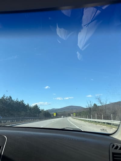 A view from inside a vehicle driving on a highway near Gorna Vasilitsa, Bulgaria. The highway is adjacent to a tree-lined area and a mountain range in the background under a clear blue sky with scattered clouds. The dashboard of the vehicle is visible in the foreground.