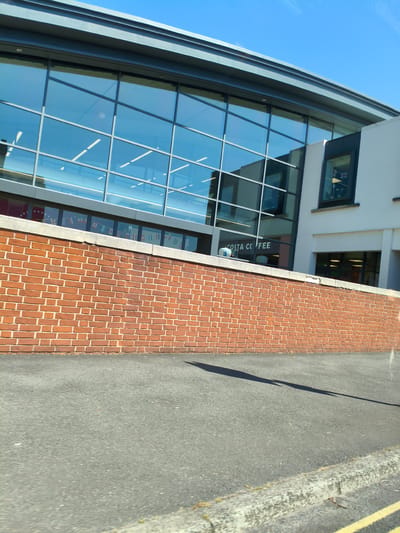The image shows a modern building with a large glass facade, reflecting the blue sky and some interior lights. Below the glass facade is a brick wall, and in front of that is an asphalt road. The building appears to be a commercial establishment, as indicated by the "COSTA COFFEE" sign visible through the glass. A small window on the right side of the building shows a flag, possibly the Union Jack. The scene is set on a sunny day. The location is Ludlow, United Kingdom.
