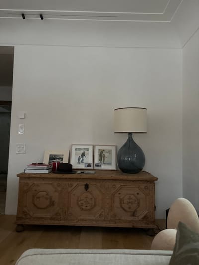 The image shows a wooden chest with intricately carved details, situated in front of a white wall with a single lamp. The chest is positioned in a room with a white ceiling featuring two black track lights.  The lamp has a cream-colored cylindrical shade with a brown trim and a dark blue glass base. On top of the chest are three framed pictures depicting people in wintery scenes. There are two white light switches on the wall. There is a hat on top of the chest and several books stacked behind it. The room also includes the partial view of a cream-colored sofa with a white cushion and a green pillow. The floor is wooden and the wall is painted white. The room is located in Celerina/Schlarigna, Switzerland.  The coordinates provided are 46.514 latitude and 9.859 longitude.