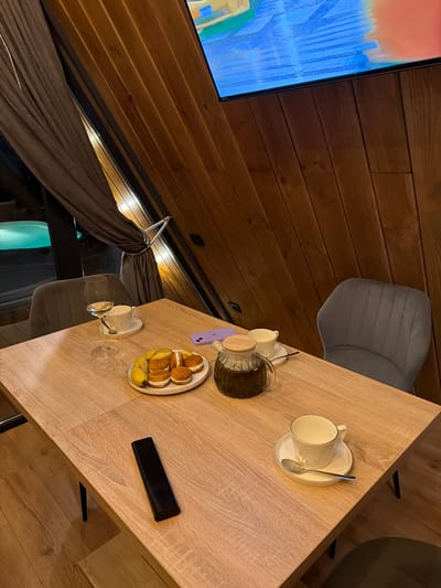 A table for two is set in a room with wood-paneled walls, possibly in a resort or spa in Sharovechka, Ukraine. The table holds a plate of cookies and bananas, a glass of white wine, a teapot filled with dark liquid, and two teacups with saucers and spoons. A remote control rests on the table. Behind the table, two grey upholstered chairs are positioned. To the left, a window looks out onto a dimly lit area with what appears to be a swimming pool. A large dark curtain is drawn to the left of the window, held back by a rope. A television screen is visible at the top right of the image, displaying a colorful abstract image. The overall ambiance suggests a relaxing or intimate setting.