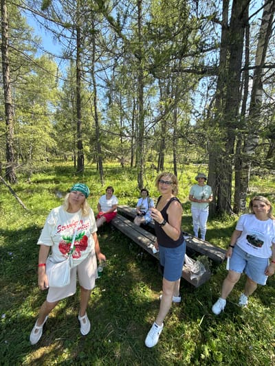 In the city of Bichiktu-Boom, Russia, a group of five women are gathered in a sunlit forest clearing on a warm day. The women are of varying ages and are dressed casually in summer attire. Two women are seated on a wooden bench, while three are standing. The woman in the foreground on the left is wearing a cream-colored t-shirt with a strawberry print, beige shorts, and a green bandana. She is holding a clear plastic bottle. To her right, another woman stands in a black and white polka dot tank top and blue shorts, holding a small sprig of greenery. In the background, three more women are visible, two of whom are also seated on the bench. One is wearing a white t-shirt and red shorts, and the other, further back, wears a white top and pants with a hat. The fifth woman is standing to the right, wearing a white t-shirt and denim shorts. The environment is characterized by tall trees with sparse foliage, a grassy ground with scattered wildflowers, and dappled sunlight filtering through the canopy. The atmosphere appears relaxed and informal, suggesting a casual outing or gathering in nature.