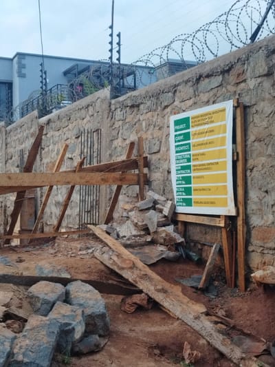 The image shows a construction site in Othaya, Kenya, located at coordinates -0.44 latitude and 36.958 longitude. The scene is set against a backdrop of a stone wall topped with barbed wire fencing. A signboard is attached to the wall, detailing information about a proposed residential development project. It includes details about the project, client, architect, structural engineer, main contractor, and various approval numbers.  The signboard is partially obscured by wooden support beams, suggesting ongoing construction. Scattered in front of the wall are numerous pieces of wood, rocks, and construction debris, indicating an active work zone. The ground is primarily brown, likely due to the presence of soil and exposed earth. The environment is characterized by an overcast sky, and a general sense of disarray, typical of a construction site.