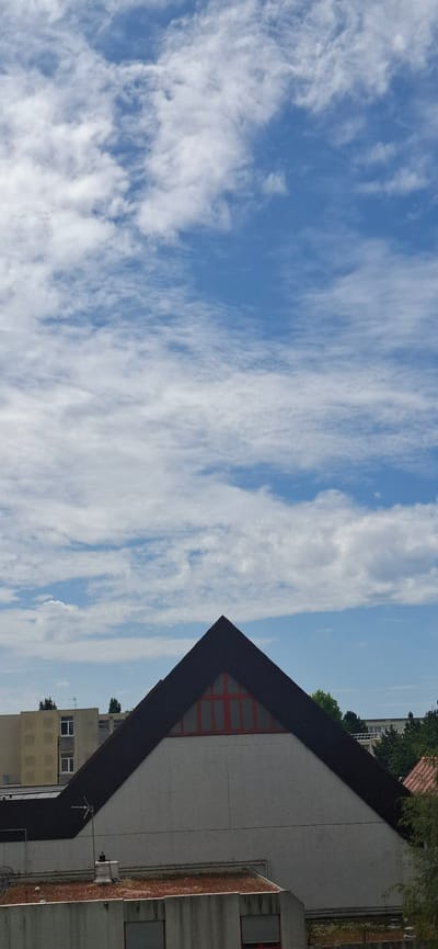 This is a photograph of a building against a bright blue sky filled with wispy white clouds. The image is oriented vertically. The building on the right has a prominent dark, angular roofline that creates a sharp, triangular shape in the frame. The main section of the building visible is a large, light-colored concrete facade with a textured appearance. A section of this facade features a pattern of red squares, suggesting windows or decorative elements. To the right of this main section, another part of the building is visible, constructed with reddish-brown brickwork, also with windows.
Further buildings can be seen in the background, taller structures with multiple windows and some greenery. The overall impression is of an urban or semi-urban environment. The bright sky and scattered clouds suggest it is daytime, likely during pleasant weather. There is no discernible activity or people in the frame. The image composition with the large expanse of sky and the sharp architectural lines of the building is visually striking. There is no visible text in the image. The location context provided is Le Havre, France.