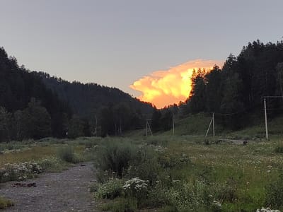 This is a horizontal, medium-distance shot of a rural landscape in Uznezya, Russia, at dusk. The scene depicts rolling hills covered in dense, dark green conifer trees on either side of a grassy, wildflower-filled meadow. A dirt path leads through the meadow from the lower left foreground. In the distance, a large, fluffy cloud is dramatically illuminated by the setting sun, glowing with vibrant shades of orange and yellow. The sky above is a pale, clear blue, transitioning to a lighter hue near the horizon. The overall mood is peaceful and serene, capturing the beauty of a rural sunset. There are no people visible, and the only man-made objects are a couple of utility poles with wires in the background, hinting at a remote settlement. The time of day is clearly indicated by the golden light on the clouds and the dimming light on the landscape.
