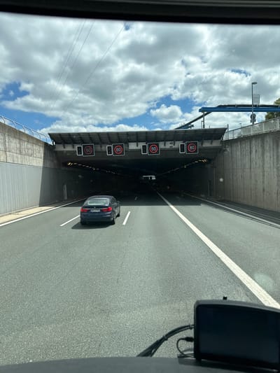 The image shows a dark blue sedan driving on a highway towards a tunnel entrance. Above the tunnel entrance, there are multiple digital signs displaying a speed limit of 80. The tunnel is lit, and it is daytime with a cloudy sky. The highway has multiple lanes, and the car is in the right lane. The photo appears to be taken from the dashboard of another vehicle. Based on the visible speed limit signs and the architecture of the tunnel and surrounding infrastructure, this location is likely the motorway A9 at the tunnel "Siechen" south of Bayreuth, Germany.