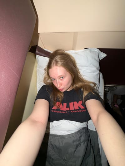 A young woman with long, light brown hair is depicted lying on her back, looking directly at the camera, likely taking a selfie. She is wearing a black t-shirt with partially visible red text that appears to include "ILK" and a chain-link design. She rests her head on a white pillow, with a dark grey or black blanket covering her lower body. Her arm is extended towards the camera, suggesting she is holding the device.

In the background, another person is partially visible, seated or reclined, with dark clothing and the letters "OK" visible on their shirt. This individual is holding a small pink object and is near a plastic water bottle. The scene is set indoors, likely a bedroom or hotel room, characterized by light beige walls and a darker background area behind the second person. A dark brown or maroon padded surface is prominent in the foreground.

The lighting is dim, indicating an evening or nighttime setting. The woman's expression is neutral and direct. The image captures a relaxed, informal moment indoors in Bobrov, Russia.