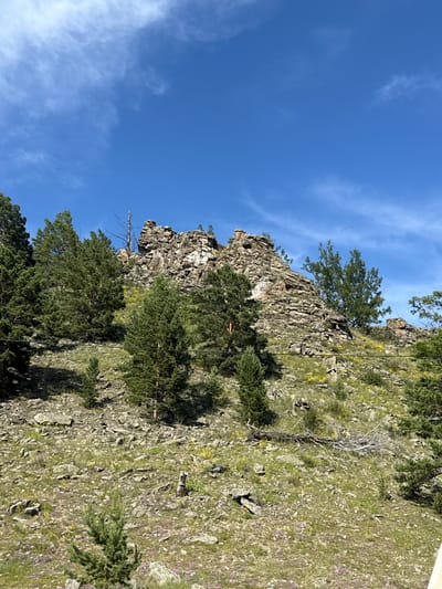 A landscape image shows a rocky hillside with sparse vegetation under a bright blue sky with wispy white clouds. The hillside is covered in rocks, grasses, and scattered pine trees. A prominent rock formation is visible at the top of the hill, with a dead tree standing near it. The scene is natural and undeveloped, suggesting a rural or wilderness area. The location is the city of Kuyada, Russia.