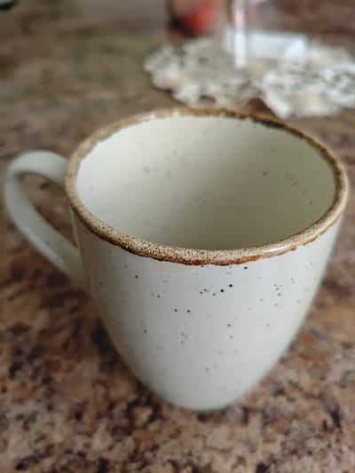 A close-up, high-angle shot shows a speckled ceramic mug with a brown rim, placed on a speckled brown countertop. The mug is white with small black speckles. The rim is a textured brown. A white handle is visible on the left side of the mug. The background is out of focus, showing a blurred brown surface and a portion of a white, lacy doily. The scene is indoors. The location is not specified as Skidzieĺ, Belarus.