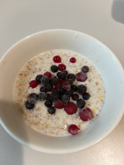 A close-up, overhead shot captures a white bowl filled with oatmeal and topped with assorted frozen berries. The oatmeal has a speckled appearance, indicating it's likely made with grains like oats and possibly other seeds. The berries include dark, frozen blueberries, some of which have a powdery frost coating, and a few red currants. A few of the berries appear slightly thawed, with small droplets of liquid around them. The bowl is positioned on a neutral-toned, possibly laminate or tabletop surface, and the background is a plain, light gray. The lighting is even, creating minimal shadows. The scene depicts a simple breakfast or snack, with the frozen berries suggesting they were recently removed from a freezer. The image does not provide any specific details about the location in Malaya Vishera, Russia, as it focuses solely on the food item.