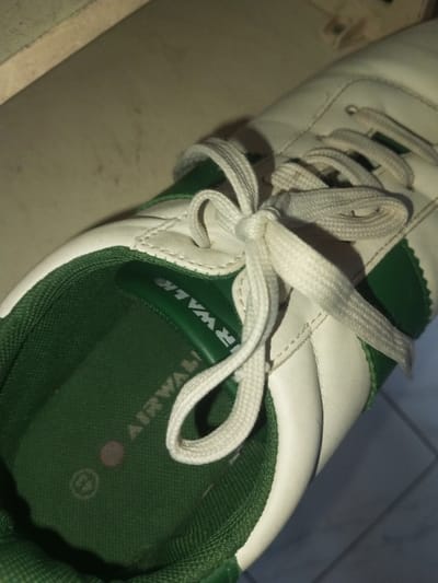 A close-up shows a white and green Airwalk sneaker with white laces tied in a knot. The interior of the shoe is green, and the brand name "AIRWALK" is visible. A small, faded pink circle is near the toe area inside the shoe. The lighting is soft, casting shadows that give the image depth.