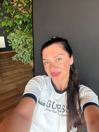 The image is a selfie of a woman in Staroabashevo, Russia. She is wearing a white t-shirt with "GUESS" and "Sports Club Los Angeles" printed on it. She has dark hair pulled back in a ponytail and is looking at the camera with a slight smile. The background is a dark wall and a wooden floor. There is also a large green plant to the left of the woman. The lighting is natural and the overall impression is a casual, indoor portrait.