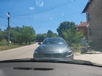 The image shows a view from inside a vehicle looking forward on a sunny day, presumably in the city of Malorad, Bulgaria. A dark-colored BMW sedan is directly ahead, parked on a paved road or driveway. To the right of the BMW, a turquoise baby stroller with large wheels is visible on a concrete surface next to a low stone wall and some vegetation. Behind the stroller and the BMW, a light-colored, multi-story building with a red-tiled roof and windows is present, along with lush green trees. To the left of the road, a wooden utility pole with multiple wires is situated amidst greenery. The sky is clear and blue. The foreground of the image is dominated by the interior of the vehicle, including the dashboard and windshield wipers.