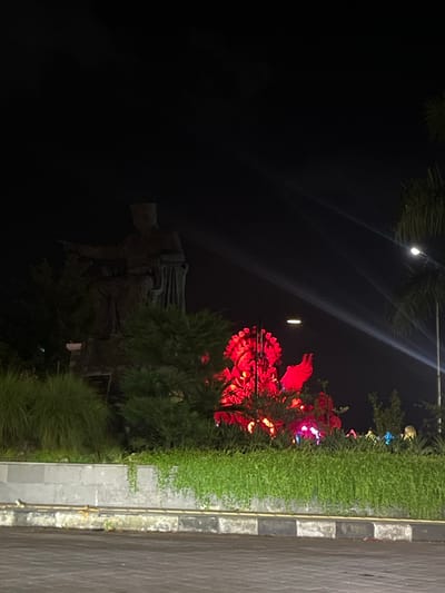 The image depicts a nighttime scene at a monument in Desa Delod Peken, Indonesia. In the foreground, a paved road surface with a black and white striped curb separates the viewer from a raised median planted with diverse green vegetation, including tall grasses and low-lying ground cover.

To the left, a dark, seated statue of a male figure is visible, wearing a cap and formal attire, with his right arm extended and index finger pointing forward. This statue is partially obscured by dark green shrubs. Behind and to the right of this figure, a large, intricate sculpture is prominently illuminated in a vibrant red light. This sculpture features multiple ornate, spiky elements and what appear to be large wings, characteristic of Balinese mythological figures.

The background is dark, indicating night, but is pierced by several strong, diagonal beams of white light originating from an unseen source on the right side of the frame, possibly spotlights. A single street light is visible on the far right, next to a partial view of a palm tree. The red light from the sculpture subtly illuminates some of the surrounding foliage. No people are visible.