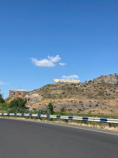 The image depicts the city of Gladzor, Armenia, with a distinctive sign on a hillside. The sign, in a stylized font with a winged emblem, reads "GLADZOR." The natural environment is arid, with dry grass covering the hills. In the foreground, a two-lane asphalt road is visible, bordered by a guardrail with alternating white and blue reflective markers. To the left of the road, on the hillside, is a brick building with a silver roof. The sky is clear and blue with a few scattered white clouds.