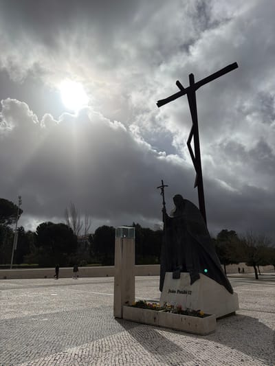 The image displays the Monument to Pope John Paul II and an accompanying large cross within the Sanctuary of Fátima, Portugal. In the foreground-midground, a dark-patinated bronze statue of Pope John Paul II stands, depicted in papal vestments, holding a crucifix in his raised right hand. The statue rests on a light-colored stone pedestal, which bears the inscription "João Paulo II." A flower box containing a small assortment of colorful blooms is positioned at the base of the pedestal. Immediately behind the statue, a tall, dark, stylized cross, constructed from what appears to be wood or metal, is erected, its form partially silhouetted. The ground is paved with light-colored, irregularly shaped cobblestones, and pronounced shadows from the monuments extend towards the viewer, indicative of strong directional light. In the mid-ground, to the left of the statue, a light-colored, rectangular pillar topped with a clear, square receptacle is visible. The background features a horizontal line of dark green coniferous trees and several lighter-toned, deciduous trees, with indistinct architectural structures partially visible beyond. Several small, dark, human figures are observed walking across the paved plaza in the mid-ground. The sky is predominantly covered by dense, dark grey clouds, with a brilliant sun breaking through a distinct clearing in the upper left quadrant, casting bright rays across the cloud cover and illuminating portions of the scene.