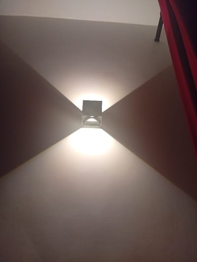A square light fixture is centered on a textured, off-white wall, emitting a bright glow that casts a symmetrical, diamond-shaped pattern of light and shadow. A section of red curtain hangs down the right side. A dark object, possibly a curtain rod, is visible at the top edge of the frame.