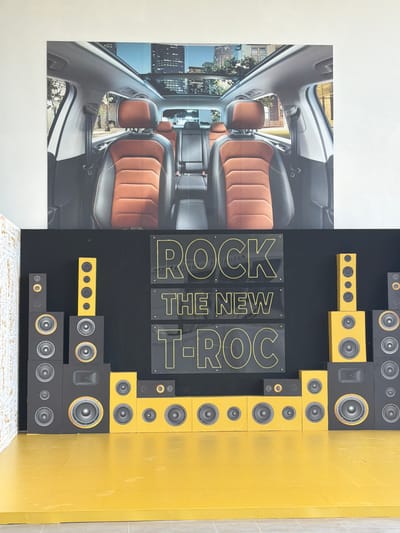 The image shows a poster advertisement with a car interior photo on the left. To the right, "ROCK THE NEW T-ROC" is displayed on individual transparent panels. A wall of speakers, some yellow and some black, borders the right side of the text. The setting's location in the city of Ouahat Sidi Brahim, Morocco, is not evident from this image.