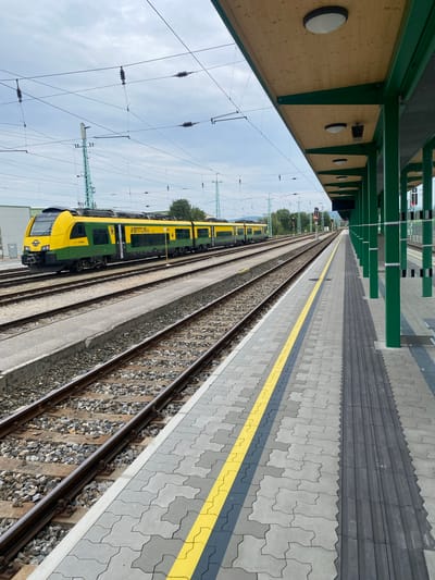 The image shows a train station platform in Wulkaprodersdorf/Vulkaprodrštof, Austria. On the left, a yellow and green train is stopped at the platform, adjacent to multiple train tracks. The train bears the inscription "VENTUS 35". The platform itself is paved with interlocking gray and yellow patterned tiles, with a tactile warning strip running along the edge closest to the tracks. To the right, a covered waiting area is supported by green pillars, with a wooden ceiling and overhead lighting fixtures. The sky is overcast with a few scattered clouds. Overhead power lines are visible across the scene, supported by tall green poles. The overall environment suggests a functional and modern train station.
