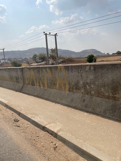 The scene depicts an outdoor landscape in Ushapa, Nigeria, under a partly cloudy sky. In the immediate foreground, a concrete highway barrier, light grey and weathered, extends horizontally across the frame. Prominently visible on the barrier's vertical face is yellow spray paint spelling "DARINA". Below the barrier, a narrow concrete sidewalk abuts a dirt and gravel road shoulder containing small pieces of debris.
Beyond the barrier, the midground features two concrete utility poles supporting multiple overhead power lines. The terrain is uneven, characterized by large exposed rock formations and sparse, dry-looking vegetation including small bushes and trees. To the far left, a cluster of residential buildings with dark roofs is visible nestled against the landscape. The background consists of gently rolling, treeless hills under a light blue sky with scattered white clouds. No people, animals, or vehicles are present.