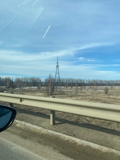 The image depicts a scene from a moving vehicle, potentially on a road adjacent to a field or open area. A guardrail runs along the right side of the frame. Beyond the guardrail, the landscape is characterized by sparse vegetation, potentially early spring or late autumn conditions. A power line tower is visible in the distance, stretching towards a blue sky with light clouds. The vehicle's side mirror is visible in the lower right corner of the frame. The location is speculatively Kueda, Russia.