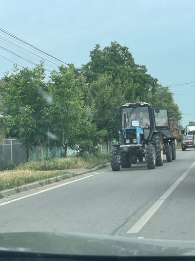 The media file depicts a blue tractor on a road in Congaz, Moldova. The tractor is the primary subject, with its large tires and distinctive blue cab being prominent. Inside the cab, a person can be seen, though their features are not clear. A sign is visible on the front of the tractor with text that reads "ЖАТВА 2020," indicating that it is participating in or related to the 2020 harvest.
The setting appears to be a rural or semi-rural area, with trees and vegetation lining the road. Power lines are also visible in the background, suggesting infrastructure present in the area. The road itself is paved with a white line marking its edge. In the distance, other vehicles, including what appears to be a truck, are present, indicating this is an active road.
The lighting suggests it is daytime, with a bright sky, though the overall impression is somewhat overcast. The image is taken from a moving vehicle, as indicated by the slight blur and the perspective. There are no strong emotional cues conveyed by the image itself, beyond the practical depiction of a working tractor in its environment. The focus is on the agricultural machinery and its immediate surroundings in the context of a harvest event.