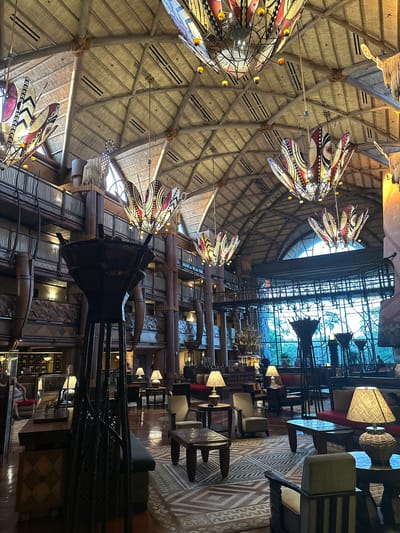 The image depicts the interior of a building located in Bay Lake, United States, showcasing a large, multi-story space with a high, thatched roof. Several large, decorative hanging fixtures with tribal-inspired designs illuminate the area. The space includes a combination of wooden beams, pillars, and various architectural details with an African or tribal aesthetic. In the background, a large window reveals glimpses of outdoor greenery. Tables and chairs are arranged on the lower levels, suggesting a dining or lounge area. A few people are visible in the image's lower portion, including a person sitting in a chair and others in the distance.