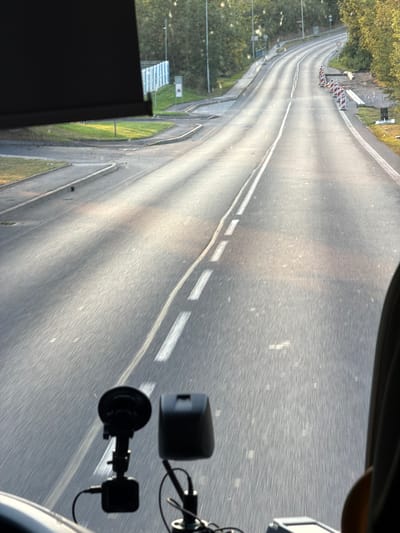 The image is a first-person perspective from inside a vehicle traveling on a two-lane road, likely in Chlumčany, Czechia. The road is asphalt, with white lane markings visible. Sunlight creates strong reflections and shadows on the road surface, indicating it's either early morning or late afternoon. On the right side of the road, there are temporary traffic barriers and some construction materials. Trees and vegetation line the sides of the road. In the foreground, two black electronic devices, possibly dashcams or GPS units, are mounted on the dashboard, with wires connecting them. Part of the vehicle's interior, including a dark sun visor and a portion of the passenger seat or curtain, is visible in the frame.