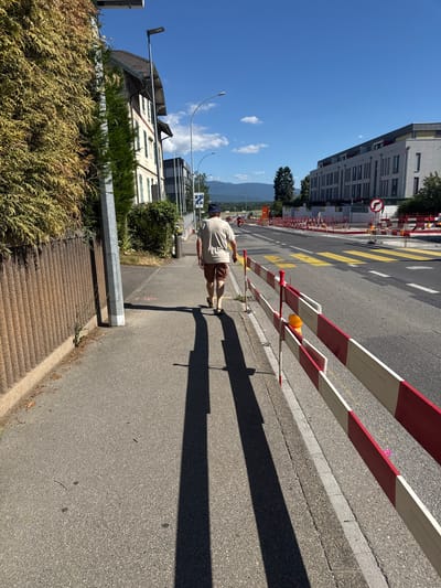 A man walks away from the camera on a paved sidewalk on a sunny day. He is wearing a light brown t-shirt and brown shorts. The sidewalk is to the left of a street that is undergoing construction, indicated by red and white barricades. Yellow pedestrian crossing markings are visible on the street. Buildings are present on both sides of the street, with lush green trees and foliage. In the background, distant mountains are visible under a clear blue sky with scattered clouds. The scene appears to be in a suburban or semi-urban area.