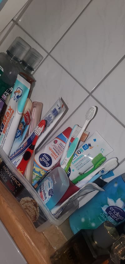 The image shows a close-up, slightly angled view of a collection of oral hygiene products and other items in a clear plastic container, resting on a wooden surface. The container is filled with various tubes of toothpaste, toothbrushes with different colored bristles, and possibly mouthwash bottles. Some visible brands and labels include "LaMax Zahncreme," "Colgate," "Donto Dent," "Junior Zahngel," "Mövenpick Cookie Dough," and "Endless Scent Laundry Granulate." The background consists of light-colored tiled walls. The lighting is somewhat dim, casting shadows and highlighting the textures of the objects. There are no people present in the image. The scene appears to be a bathroom or a similar personal care area. The image does not contain any identifiable landmarks or contextual clues that would suggest a specific location within Sasbach, Germany.