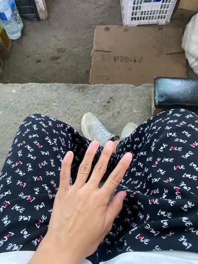 The image shows a close-up view of a person's legs wearing black pants with a repeating pattern of the word "LOVE" in white and red hearts. A person's hand rests on their left thigh, fingers splayed. The person is wearing light gray sneakers. The background is blurred but appears to be an outdoor setting, possibly a street or sidewalk. There are some objects in the background, including a white plastic crate, a cardboard box, and a white bag. The lighting suggests it is daytime. The exact location in Kyzyl-Kyya, Kyrgyzstan, cannot be determined from the image alone.