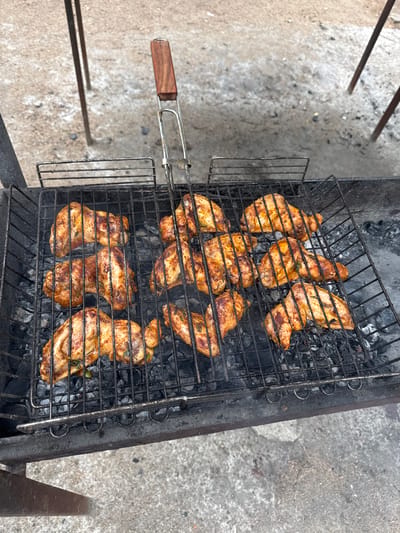 In Aiyrtau, Kazakhstan, an outdoor barbecue is underway. Chicken wings are arranged on a metal grill rack, some with visible herbs. The grill is positioned over hot coals within a dark metal barbecue pit. The background shows a gravelly ground surface and the legs of a tripod-like structure. A wooden-handled metal tool, likely part of the grill mechanism, extends from the top of the grill. The scene suggests a casual outdoor cooking event.