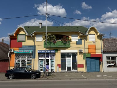 The image shows a multi-story building with a colorful facade in Sremska Mitrovica, Serbia. The building has a green roof and features various geometric shapes in different colors on its exterior, including red, yellow, orange, and blue. Several windows with white roller shutters are visible on the upper floors, and there is a balcony with flowering plants.
On the ground floor, there are two businesses: "STOLARSKI CENTAR" (Carpentry Center) and "STOLARIJA MLADENOVIĆ" (Mladenović Carpentry). The "STOLARSKI CENTAR" has a rolling metal gate and displays advertising inside. The "STOLARIJA MLADENOVIĆ" has a glass door and displays information about their services.
A black sedan is parked in front of the building, and a bicycle is leaning against a pole on the sidewalk. A street light pole with wires runs across the top of the image. The sky is blue with scattered white clouds. The building is situated on a street with a paved sidewalk and road. To the right of the colorful building, a portion of an adjacent building with a tiled roof and a window displaying items is visible. The image appears to be taken on a sunny day.