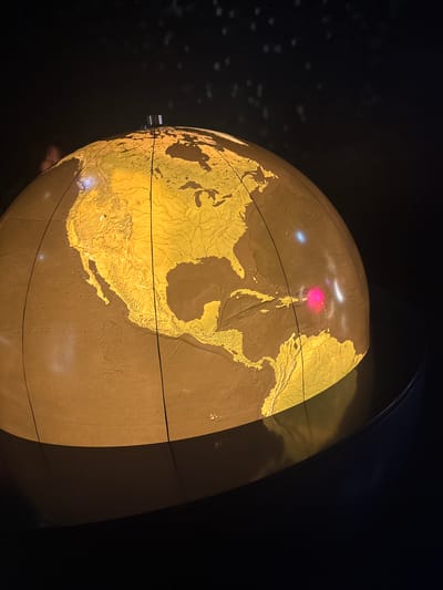 The image depicts a globe with illuminated continents against a dark background. The land masses are brown and yellow, with visible details of rivers and mountain ranges. A horizontal line encircles the globe, likely representing the equator. There are reflections on the globe's surface, including a red light. The globe appears to be cut in half, revealing the interior structure or another part of the map. There is a metallic element attached to the left side of the globe. Given the context, the globe is most likely an exhibit in the Lindt Home of Chocolate Museum located in Kilchberg (ZH), Switzerland.