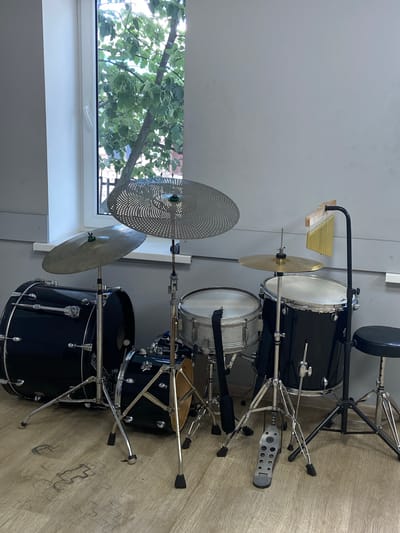 A drum set is situated in a room with a large window. The drum set is comprised of various drums, cymbals, and a bass drum pedal. The drums are black and silver. A black stool is positioned to the right of the drum set. The room has light gray walls and a wooden floor with scuff marks. Outside the window, a tree with green leaves is visible, along with a hint of a fence and buildings, suggesting an urban or suburban setting in Soroca, Moldova. No people are present in the image.