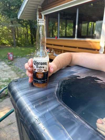 In Saue, Estonia, a person's arm rests on the edge of a hot tub, holding a bottle of Corona Extra beer. The hot tub is dark blue and filled with water, with bubbles forming on the surface. The background shows a wooden building with large windows, surrounded by trees and greenery. The scene is outdoors, suggesting a recreational setting.