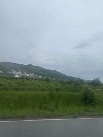 The image shows a landscape view in Mirkovo, Bulgaria, taken from a moving vehicle. A mountain range is visible in the background, covered with dense green vegetation. In the midground, there is a large industrial complex with tall structures and smokestacks. The sky is overcast and gray, suggesting a cloudy or rainy day. The foreground shows the edge of a road or highway.