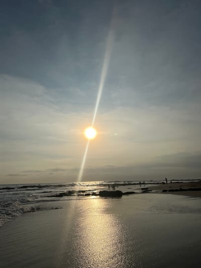 The image shows a beach scene at sunset, taken in Santa Teresa, Costa Rica, specifically at the coordinates 9.635 latitude and -85.161 longitude. The sun, partially obscured by a cloud, shines brightly in the sky and casts a long ray of light down through the clouds. The sky is a mix of light blue and white clouds. In the foreground, the beach is wet and sandy, with some rocks and small waves breaking along the shoreline. Several people are standing on the beach, silhouetted against the sun. The water is calm and reflects the sunlight, creating a sparkling effect. The horizon is in the distance and the ocean is calm and expansive.