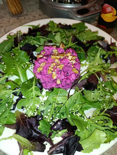 This is a close-up, eye-level, indoor shot of a salad. The salad is served on a white plate and is composed of mixed greens, including dark red and light green leaves. Scattered throughout the greens are white and black sesame seeds. In the center of the salad is a mound of bright pink food, topped with chopped walnuts. The background is blurry but appears to be a kitchen counter with a stainless steel appliance and a bottle of what looks like fruit juice. There are no people visible in the image. The Municipal Unit of Meliteieis is not depicted in this image.