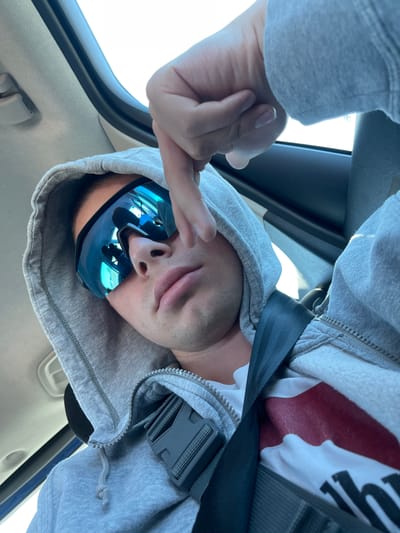 A person wearing sunglasses and a gray hooded sweatshirt is seated in a vehicle, secured with a seatbelt. The person is making a hand gesture with their middle finger extended. The interior of the car is visible, with the dashboard and window frame partially in view. The image was taken upside down. The location of Ceprano, Italy is not determinable from the image.