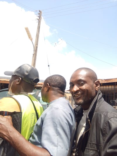 In Iseyin, Nigeria, three men are pictured outdoors on a sunny day, with a clear sky and some clouds visible above. The man in the foreground on the right, facing the camera, has a shaved head, dark skin, and a beard. He is smiling and wearing a black leather jacket over a white t-shirt. To his left, a second man, with dark skin and a shaved head, is seen from the back, wearing a light blue tunic. The third man, also seen from the back and the left, wears a black baseball cap and a bright yellow reflective vest over an orange shirt. He is wearing clear glasses. Behind the men, a wooden utility pole with overhead wires and a street light is visible, along with corrugated metal roofing and building structures. A portion of a vehicle is also present. The overall impression is of a candid, outdoor moment.
